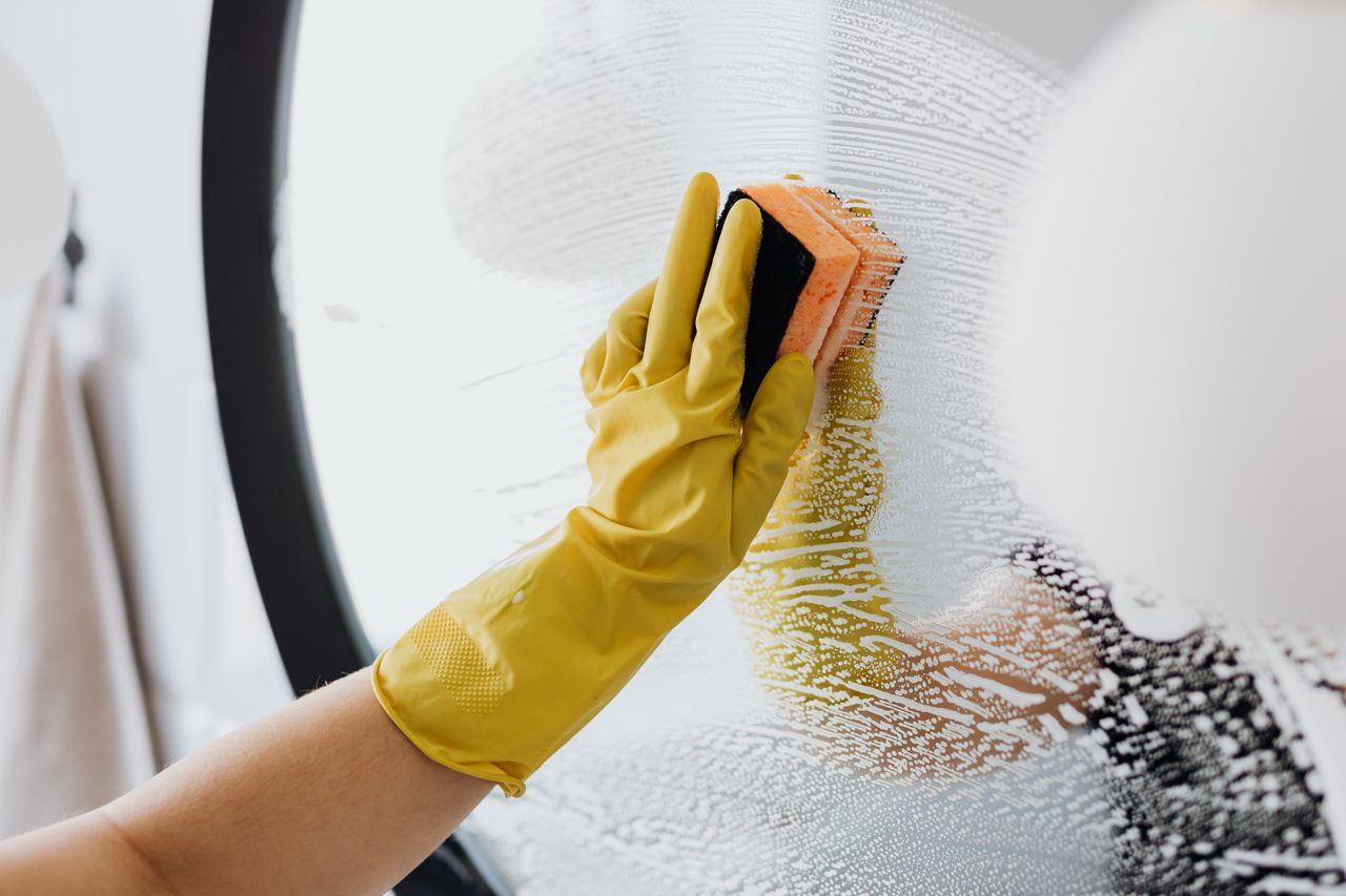 Anonymous person in yellow rubber gloves using a plain sponge to clean a bathroom mirror